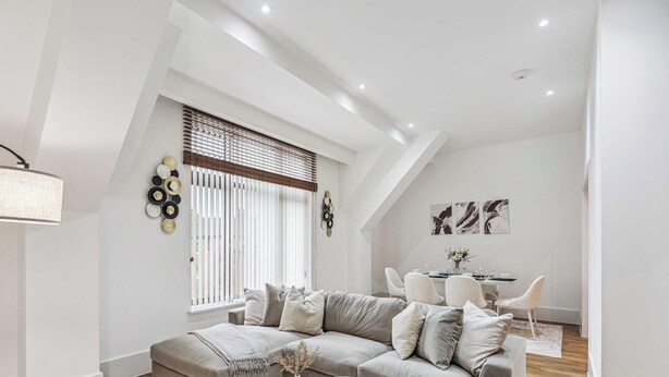 Een moderne woonkamer met een bank, een eethoek met tafel en stoelen, en een groot raam met jaloezieën.