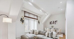 A modern living room with a sofa, a dining area with a table and chairs, and a large window with blinds. - The Hamilton Suite - 2Bed Apartment with Parking (Henley-on-Thames)