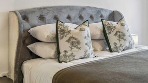 Een bed met een gestoffeerd hoofdeinde, meerdere kussens met bloemenmotieven en een decoratief object aan de muur.