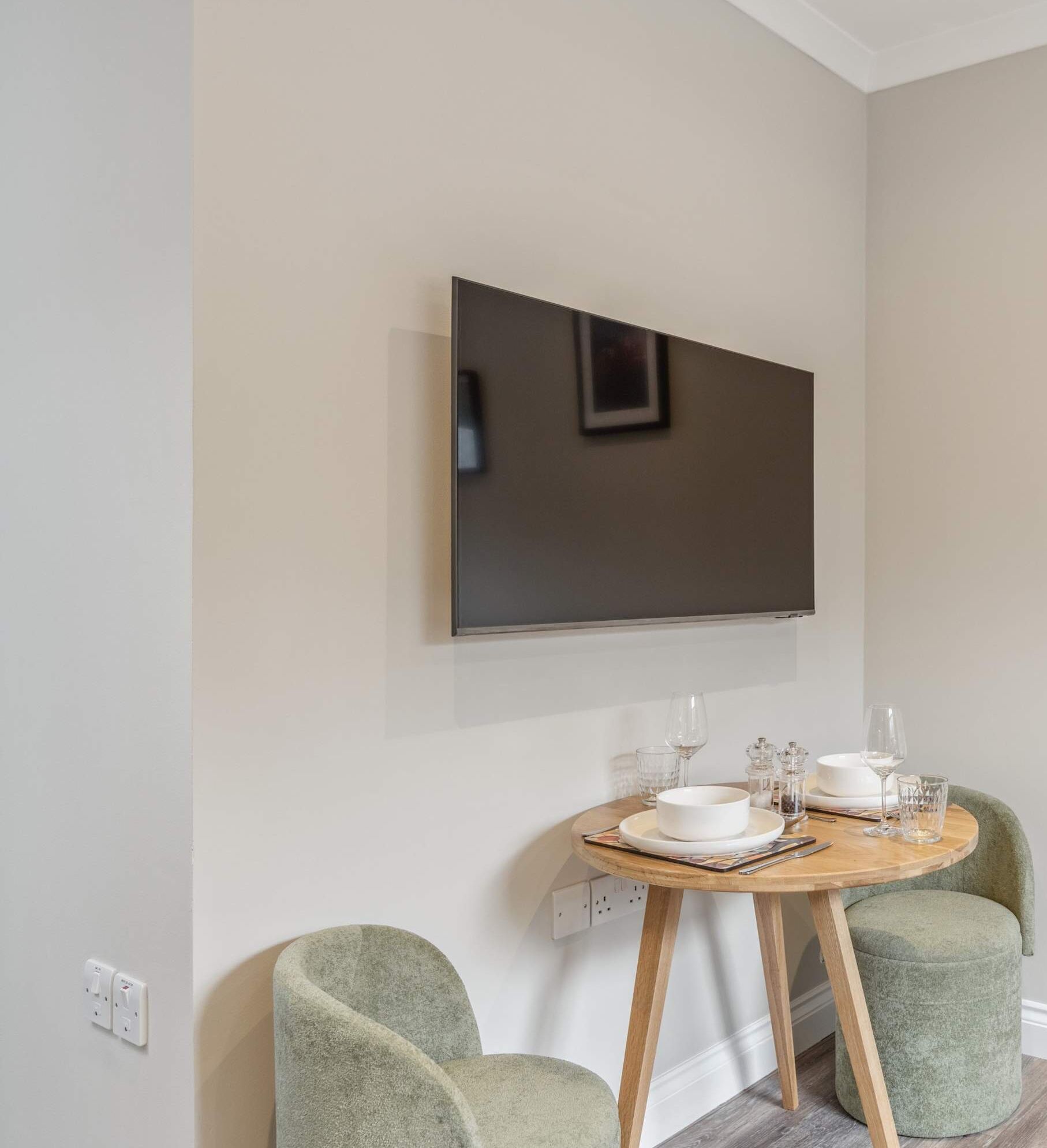 A modern dining area with a round wooden table, two green upholstered chairs, a flat-screen TV mounted on the wall, and a small round ottoman.