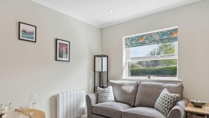 Un salon moderne avec un canapé gris, deux tableaux encadrés, une lampe sur pied et une fenêtre donnant sur un paysage verdoyant.