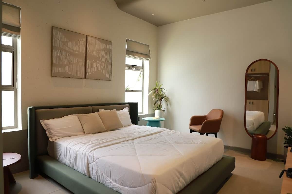 A bedroom with a large bed, a chair, a small table, a mirror, and a potted plant.