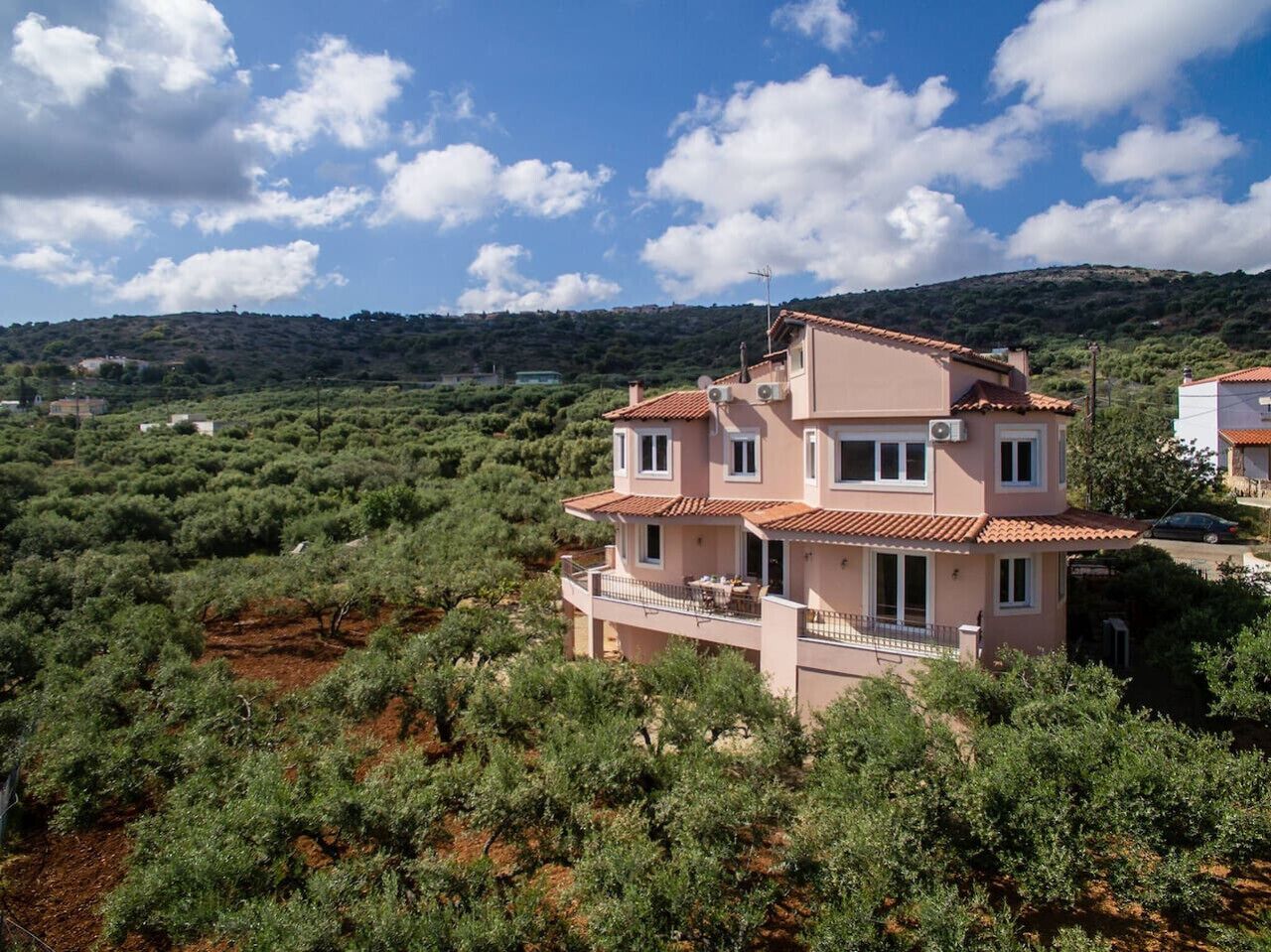 A two-story house with a balcony, surrounded by olive trees and a hilly landscape.