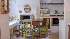 A cozy dining area with a round wooden table, green chairs, and a cowhide rug. A fireplace with a decorative mirror and a hanging light fixture is visible. - Can Sabuba (Deia)