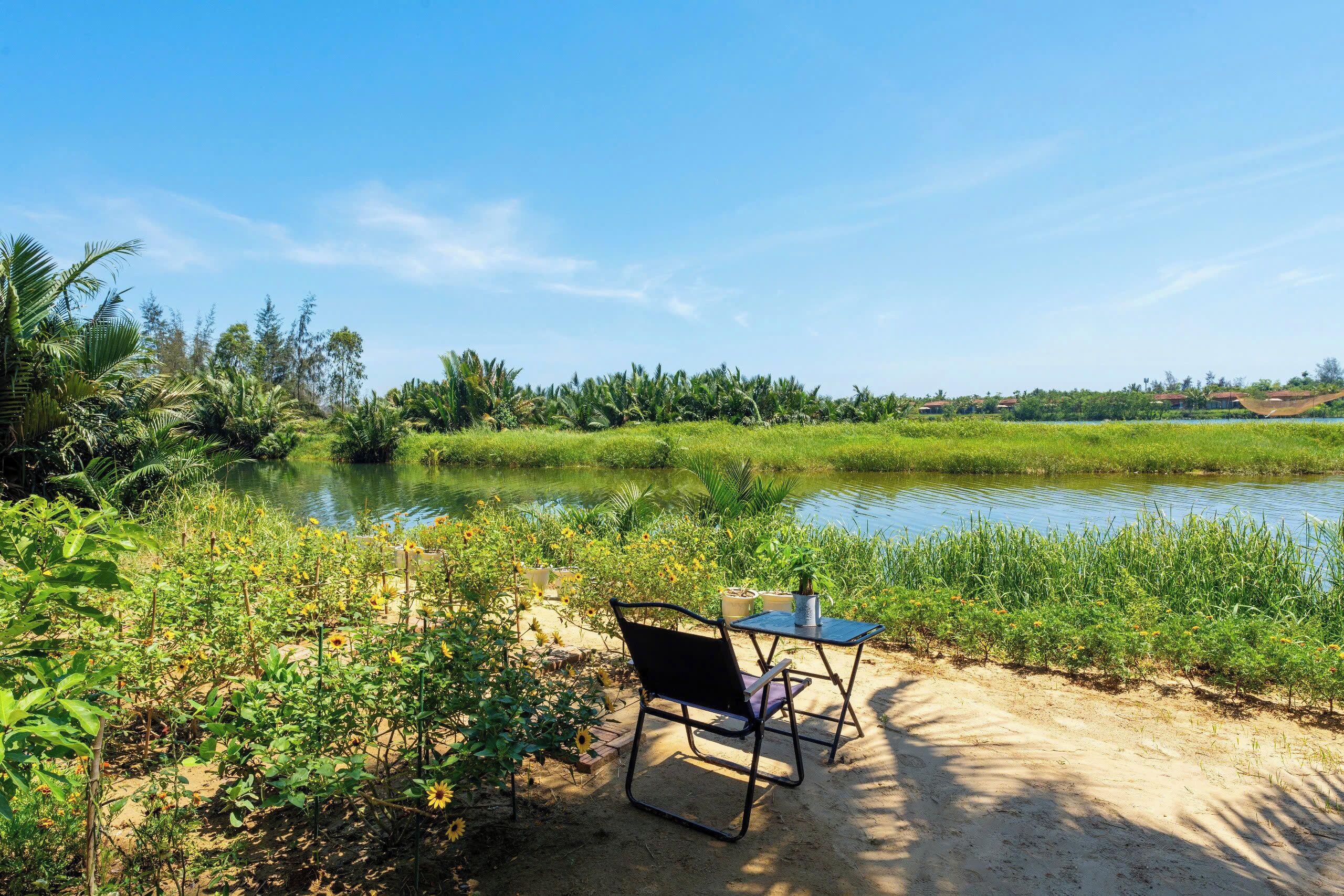 A chair and table by a water body with greenery and distant buildings.