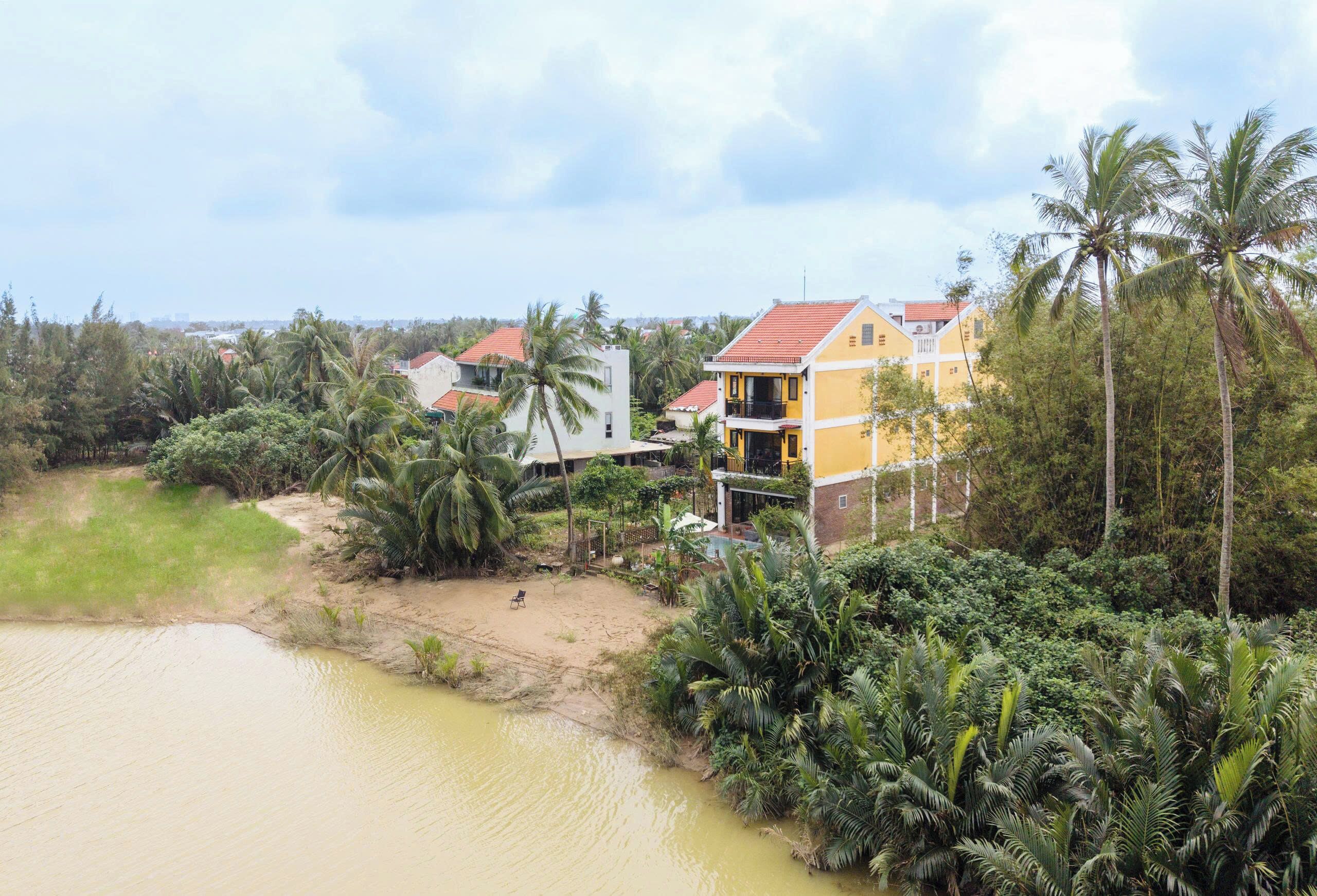 A riverside property with a yellow building, palm trees, and a sandy beach.
