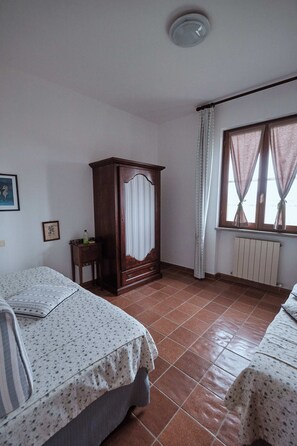 A bedroom with two beds, a wooden wardrobe, a radiator, and a window with curtains. - Belvedere Apartment - Peacock Farmhouse (Castiglione della Pescaia)