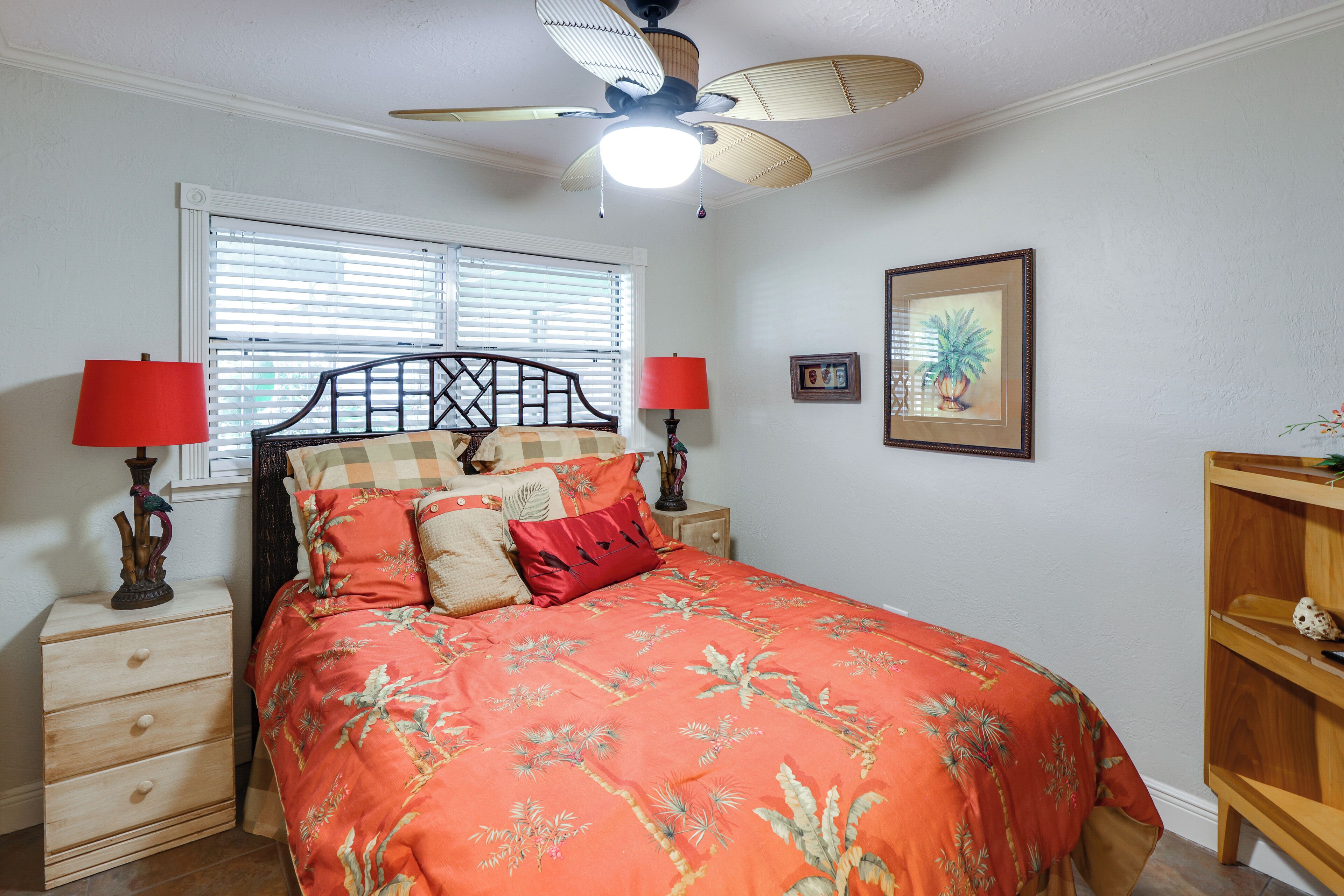 A bedroom with a bed, two nightstands, a ceiling fan, a window with blinds, and a framed picture on the wall.