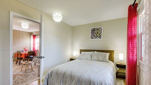 A bedroom with a large bed, bedside tables, a nightstand, a red curtain, a painting on the wall, and a dining area visible through an open door. - Convenient + Cozy! Apartment in DC Suburbs! (Silver Spring)