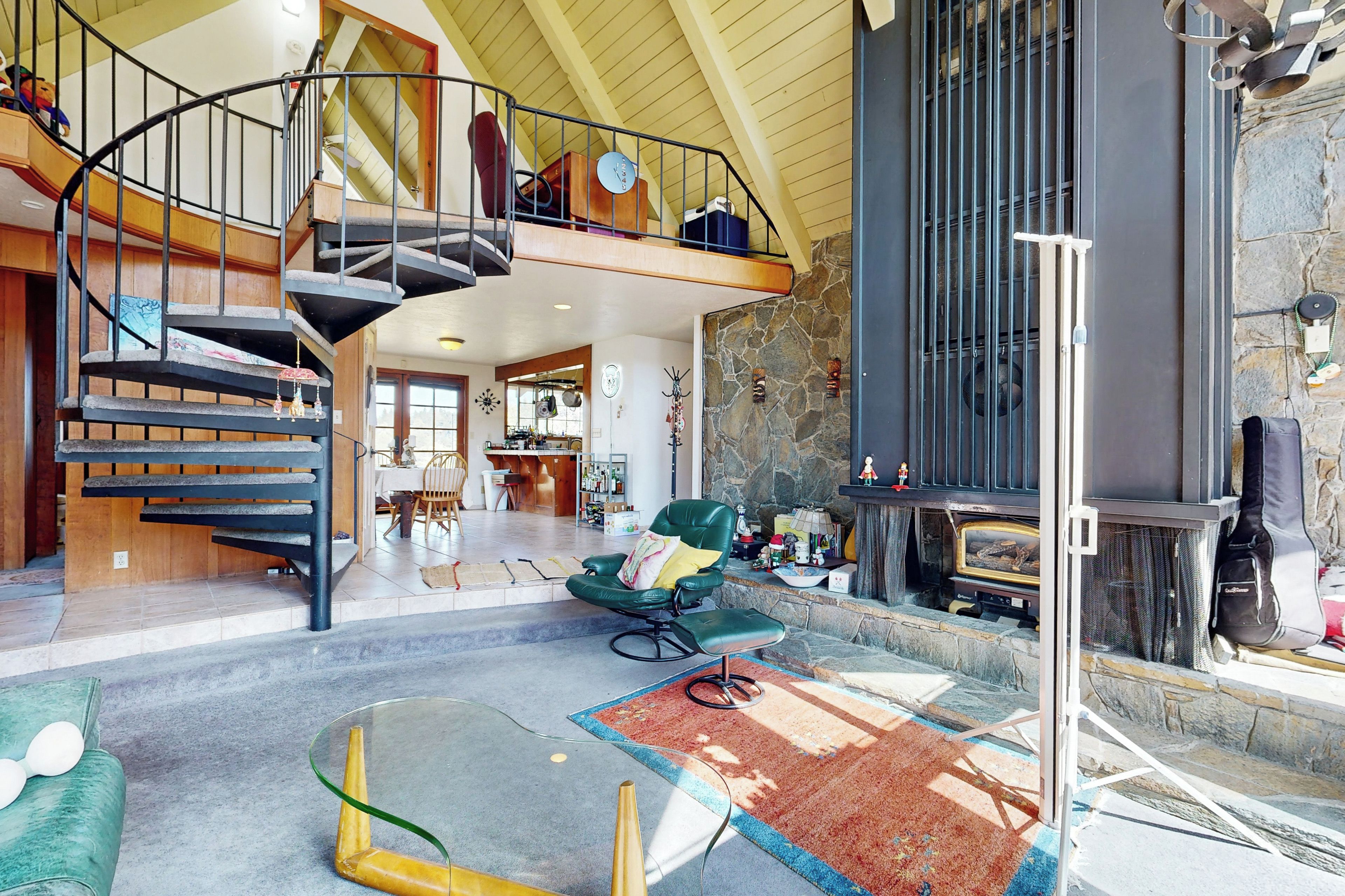 A spacious living area with a high ceiling, a stone fireplace, a green leather chair, a glass coffee table, and a spiral staircase leading to an upper level.