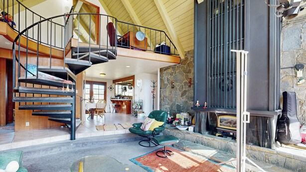 Amplia sala de estar con techo alto, chimenea de piedra, un sillón de cuero verde, una mesa de centro de vidrio y una escalera en espiral que conduce a un nivel superior.