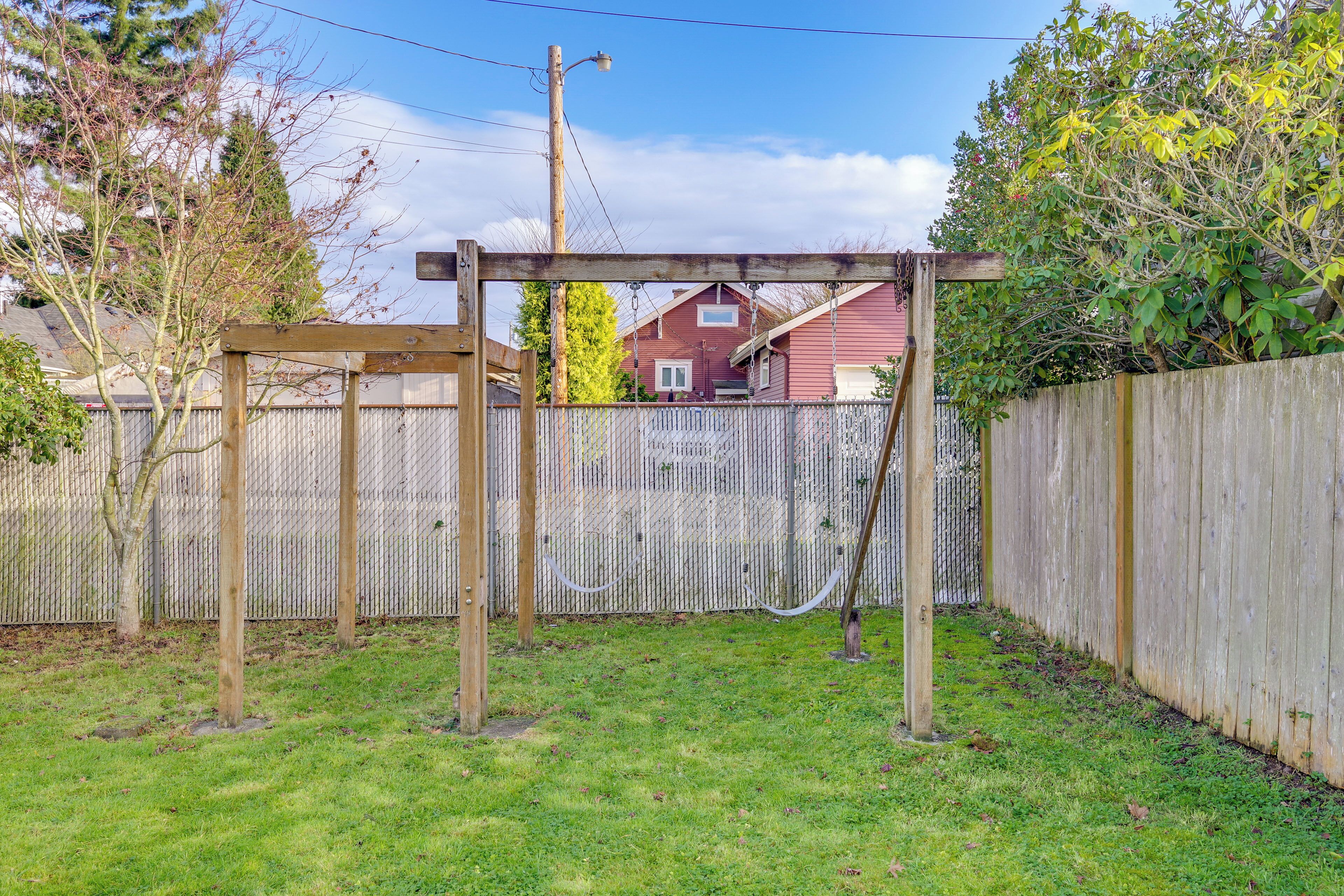 Ein hölzernes Schaukelgestell in einem Garten mit einem Maschendrahtzaun, einer Holzterrasse und einem roten Haus im Hintergrund.