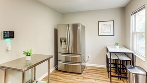 Une cuisine compacte avec un réfrigérateur en acier inoxydable, une petite table à manger avec des chaises, une plante en pot et un tableau encadré au mur.