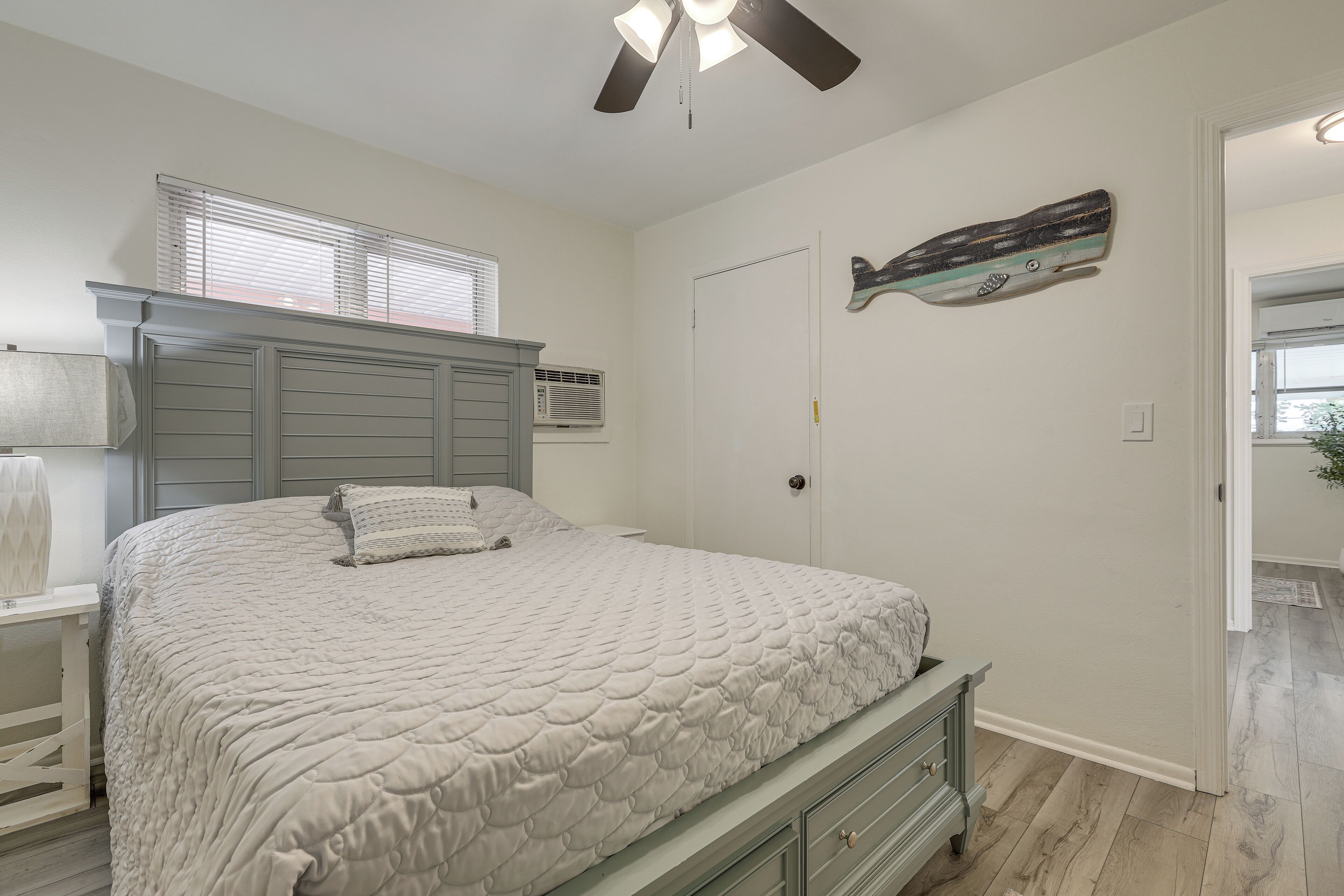 Une chambre à coucher comprenant un lit, une commode, un ventilateur de plafond, une porte et une fenêtre avec des stores.