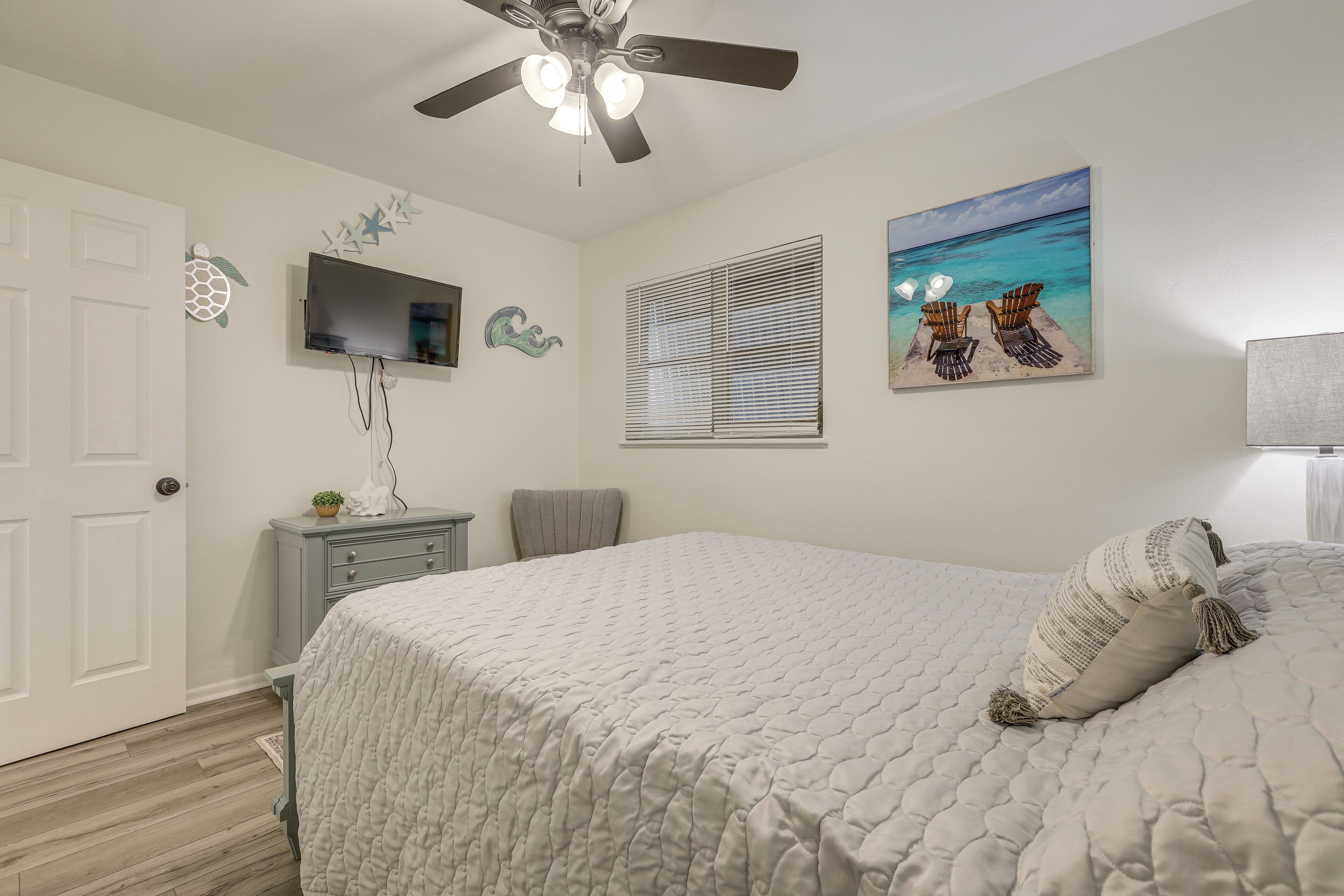 A bedroom with a bed, a ceiling fan, a TV, a window with blinds, and a beach-themed painting.