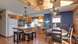 Cocina con armarios de madera, una isla central con taburetes, una mesa de comedor con sillas e iluminación colgante.