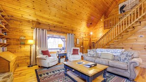A cabin interior with a wooden staircase, a sofa, armchairs, a coffee table, and a rug. - Creek On-site! Cabin 7 Mi to Cataloochee Ski Area (Maggie Valley)