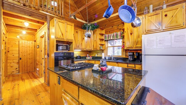 A wooden cabin kitchen with granite countertops, wooden cabinets, a stove, and hanging pots. - Creek On-site! Cabin 7 Mi to Cataloochee Ski Area (Maggie Valley)
