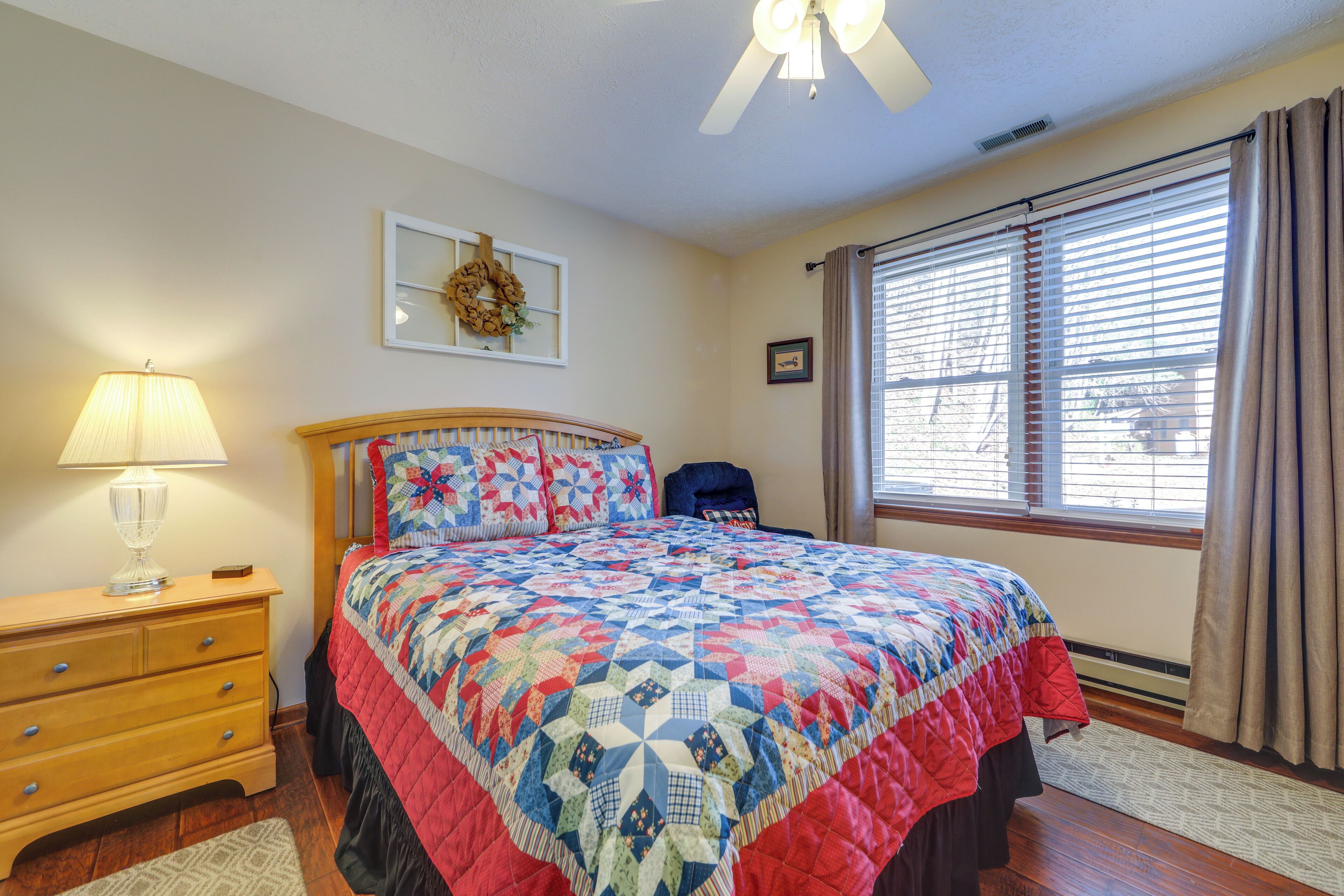 A bedroom with a wooden bed, a nightstand with a lamp, a dresser, a window with blinds, and a ceiling fan.