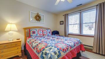 A bedroom with a wooden bed, a nightstand with a lamp, a dresser, a window with blinds, and a ceiling fan.