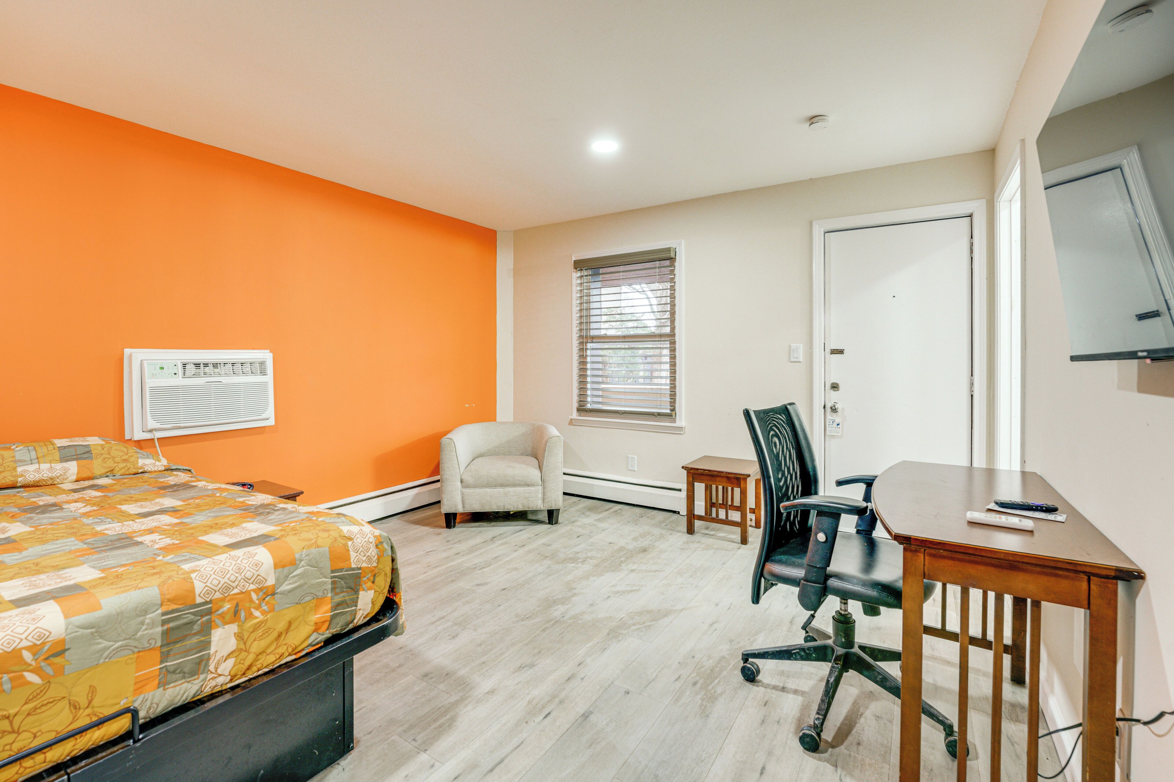A bedroom with an orange wall, a bed with a patterned blanket, a chair, a desk, and a window with blinds.