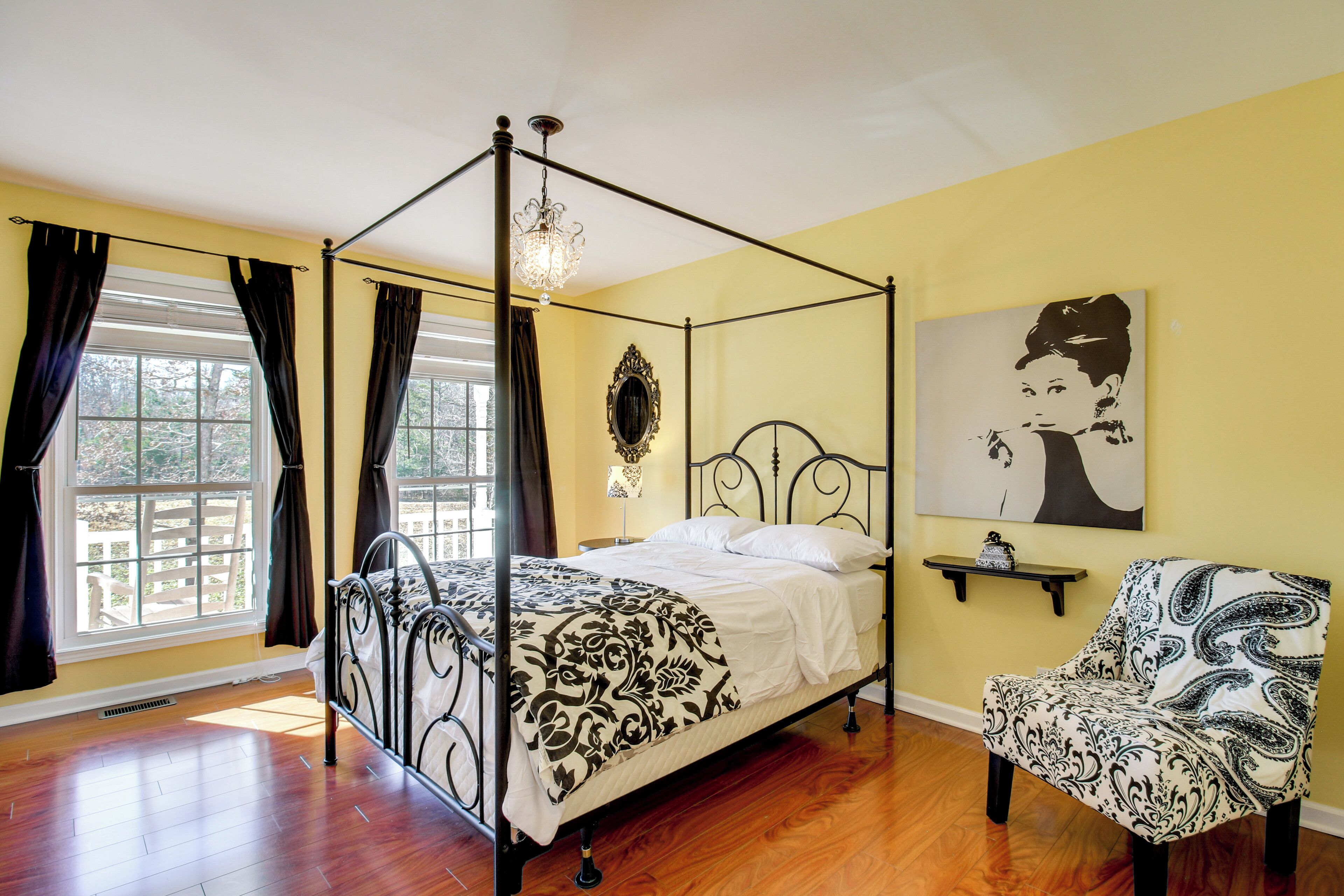 A four-poster bed with a black and white patterned bedspread, a chair with a black and white paisley design, a chandelier, and a painting on the wall.