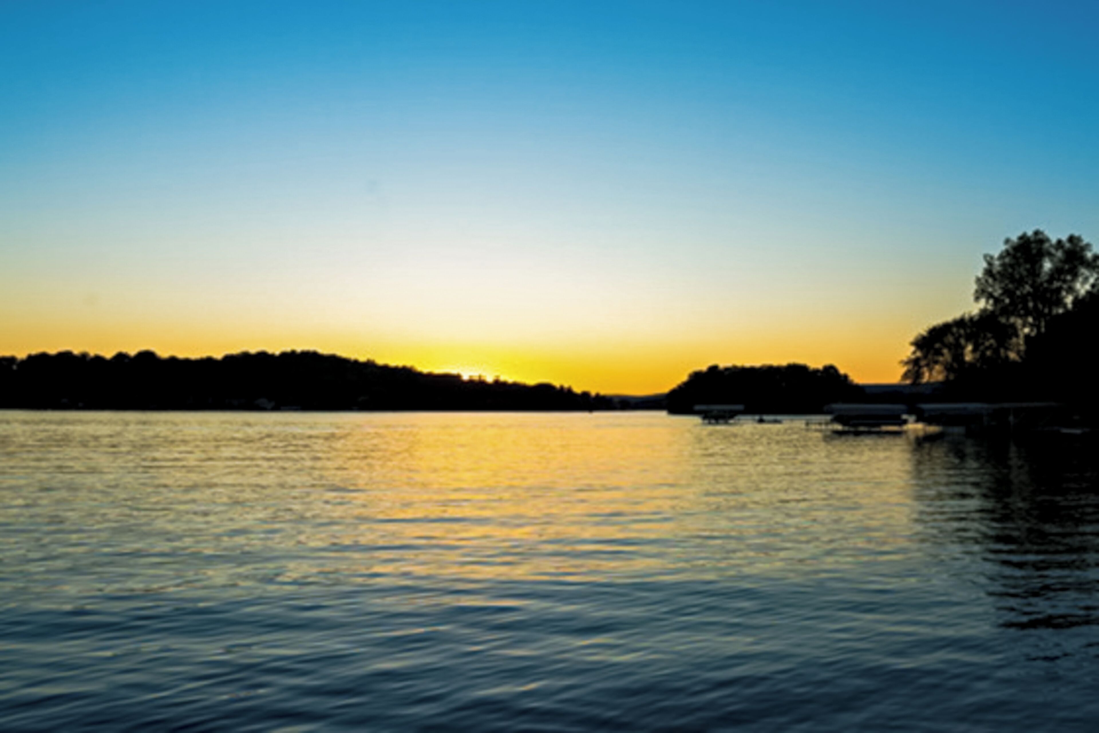 A calm river at sunset with a silhouette of trees and a dock.
