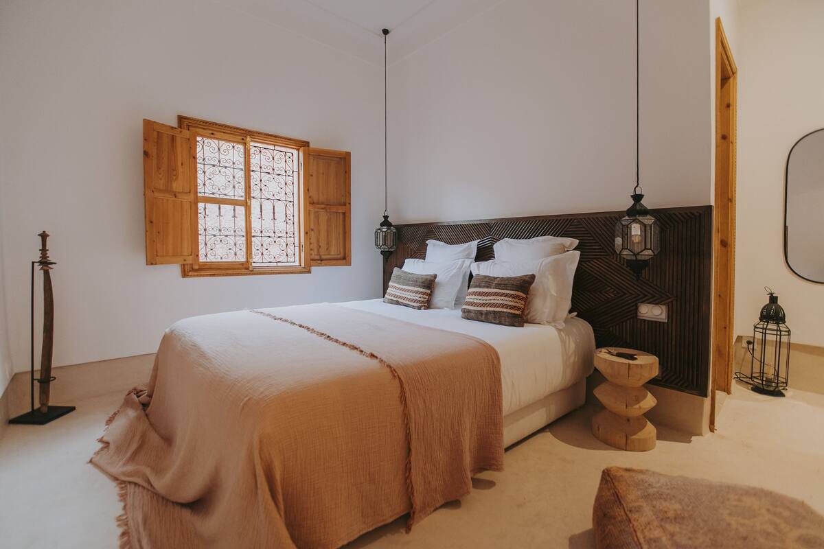A bedroom with a bed, wooden headboard, bedside lamps, a window with shutters, and a bedside table with a lantern.