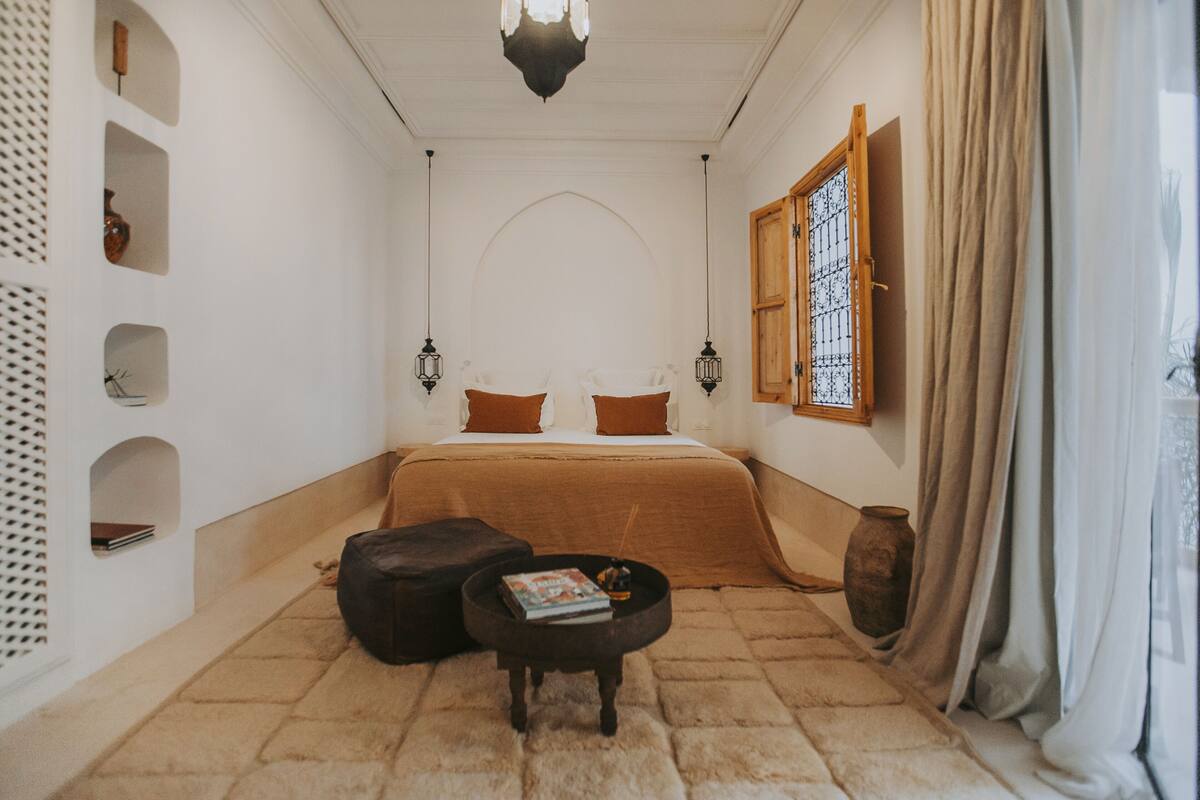 A room with a bed, a small table, a window with a wooden frame, and a wall with shelves.
