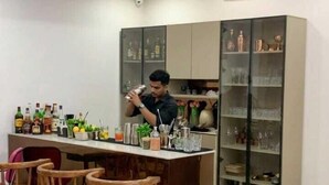 A modern kitchen with a bar area, equipped with bar stools, a countertop with various bottles, and a large cabinet with glassware. - AKD HOMES (Noida)