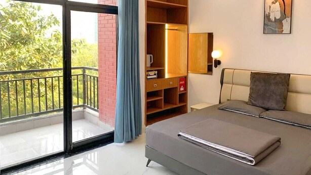 A bedroom with a bed, a wardrobe, a nightstand, a wall-mounted lamp, and a view of greenery through a balcony. - AKD HOMES (Noida)