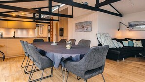 Een moderne eetruimte met een houten tafel en grijs gestoffeerde stoelen, een zwarte bank met gekleurde kussens en een keuken op de achtergrond.