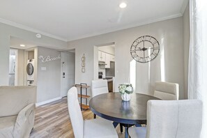 Un salon moderne avec une table à manger ronde, des canapés de couleur claire, une grande horloge murale et une cuisine en arrière-plan.