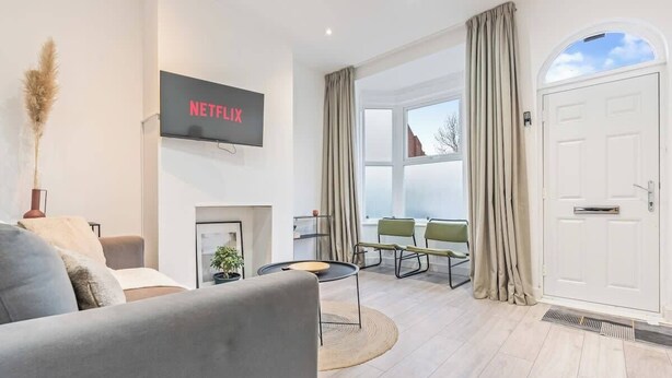 Ein modernes Wohnzimmer mit flachbildfernseher an der Wand, einer Couch, einem Couchtisch und einem Kamin. Zwei Sessel und ein Fenster mit Vorhängen sind ebenfalls vorhanden.