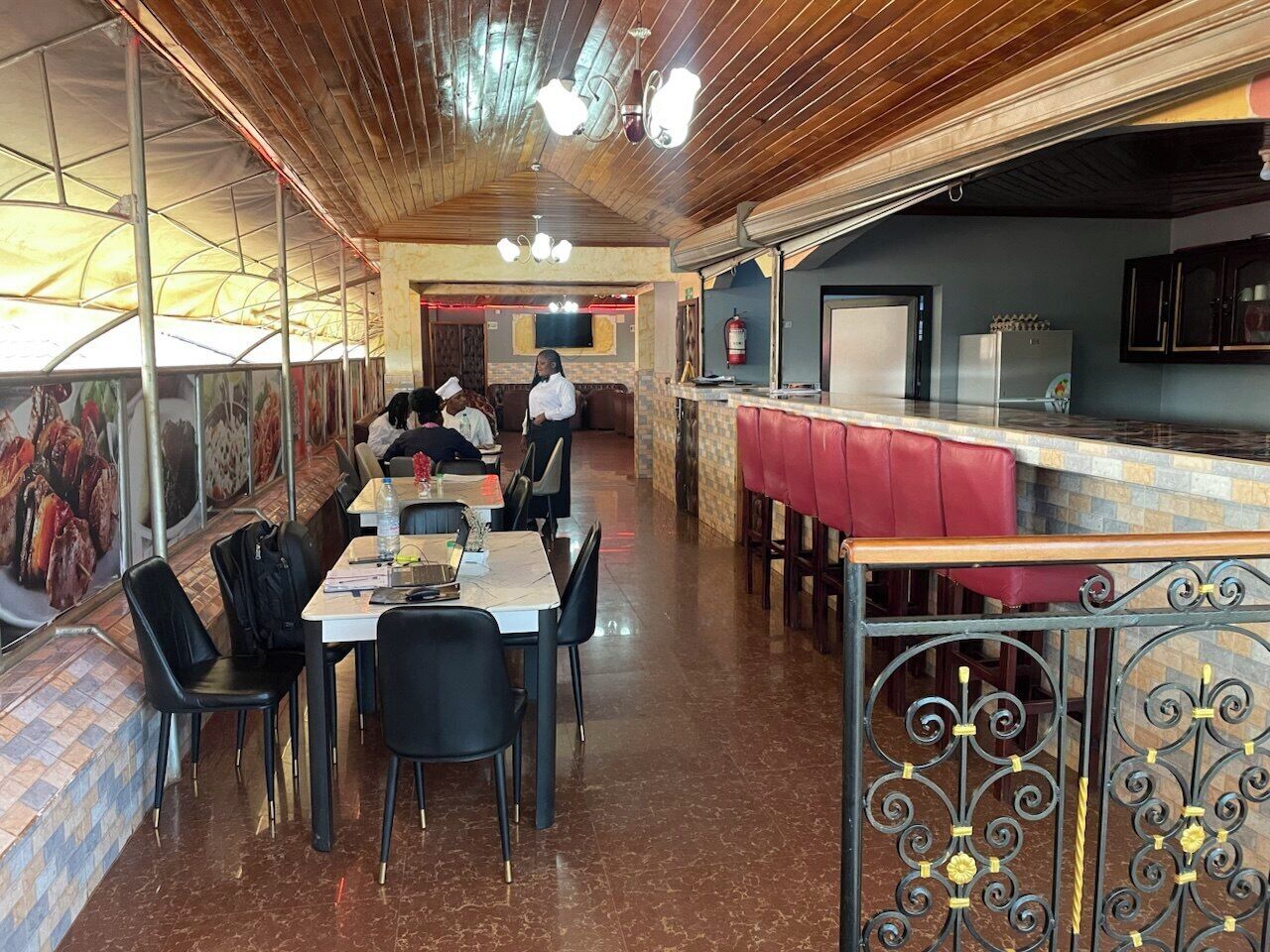 A restaurant with a long counter, multiple tables, and chairs. There are two people in the background, one standing and one seated. The ceiling has a wooden design with hanging lights.
