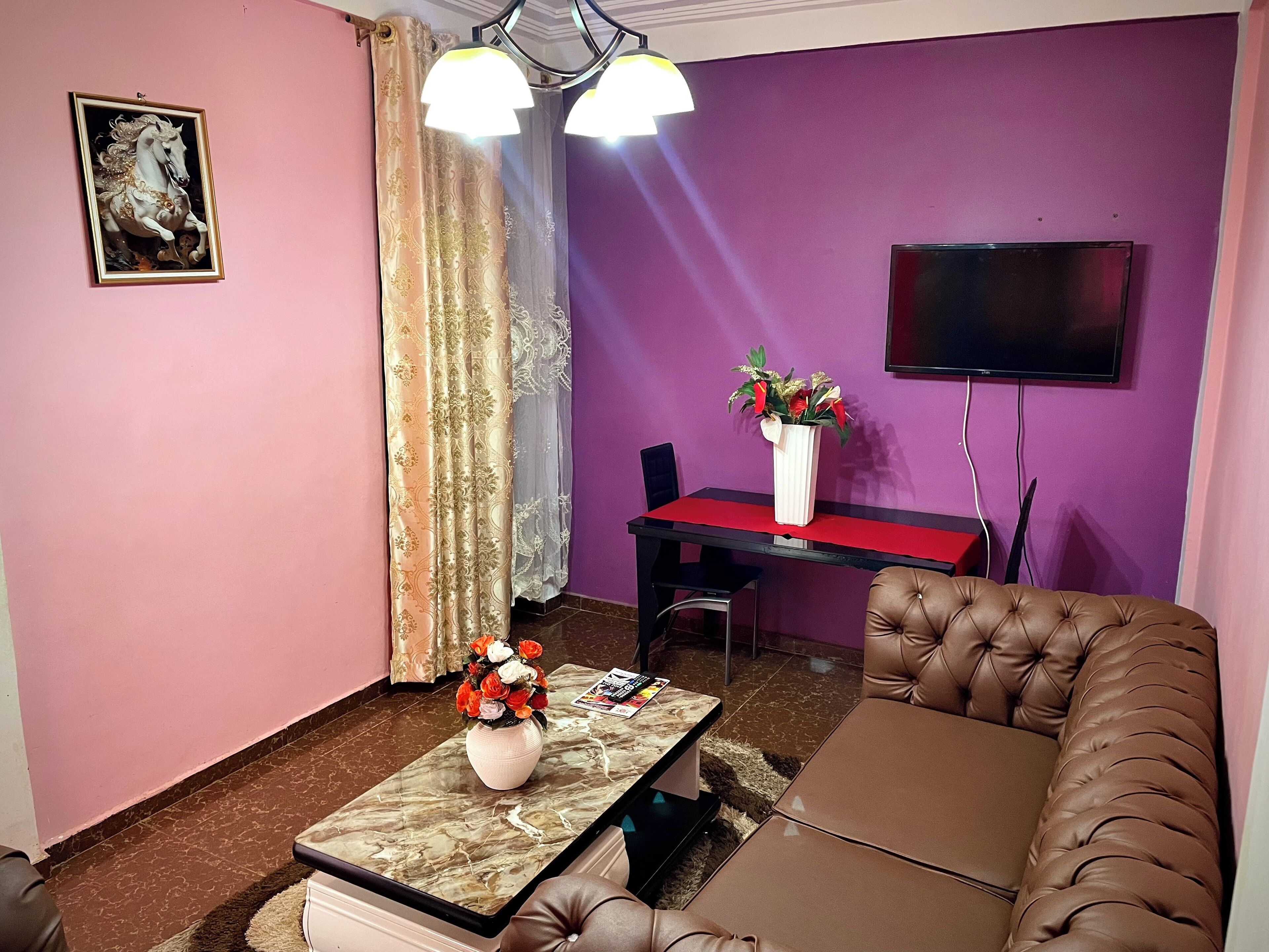 Un salon comprenant un canapé, une table basse ornée d’un vase de fleurs, un téléviseur fixé au mur et un bureau avec une chaise.