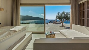 A modern hotel room with a large sliding glass door leading to a balcony with a view of the sea and mountains. There are two white sofas and a small table with a book on it. - Villa Noema - Seaside Living Redefined (Agiassos)