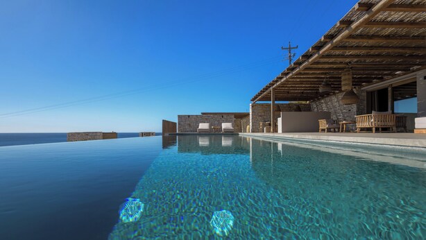 An infinity pool with clear water, a stone wall, and a wooden pergola with seating. - Villa Noema - Seaside Living Redefined (Agiassos)