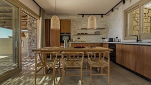A modern kitchen with a wooden table and chairs, stone walls, and hanging light fixtures. - Villa Noema - Seaside Living Redefined (Agiassos)