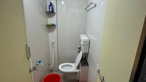 Un baño pequeño con inodoro, ducha, un cubo rojo y un taburete.
