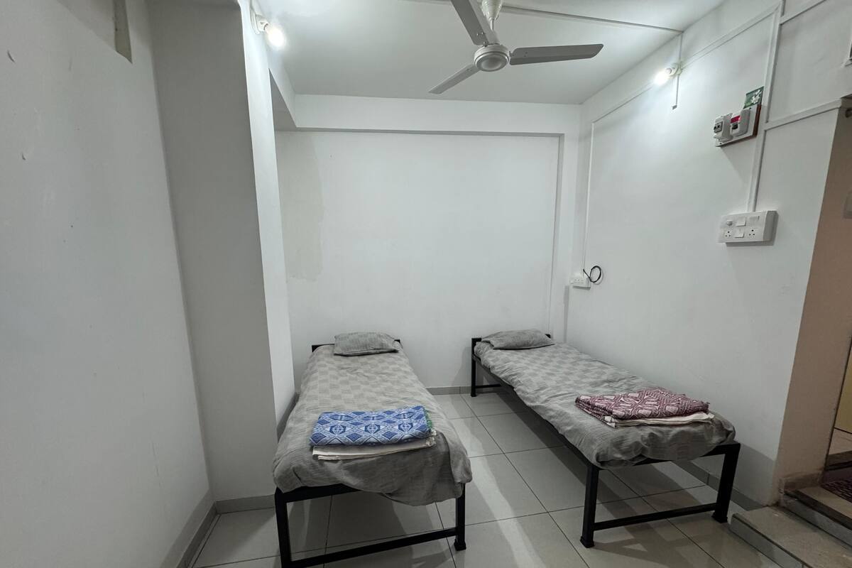 Une petite chambre propre, avec deux lits simples, un ventilateur de plafond et une prise électrique au mur.