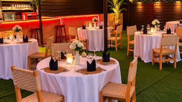 Een terras met ronde tafels, rieten stoelen en een bar met flessen.