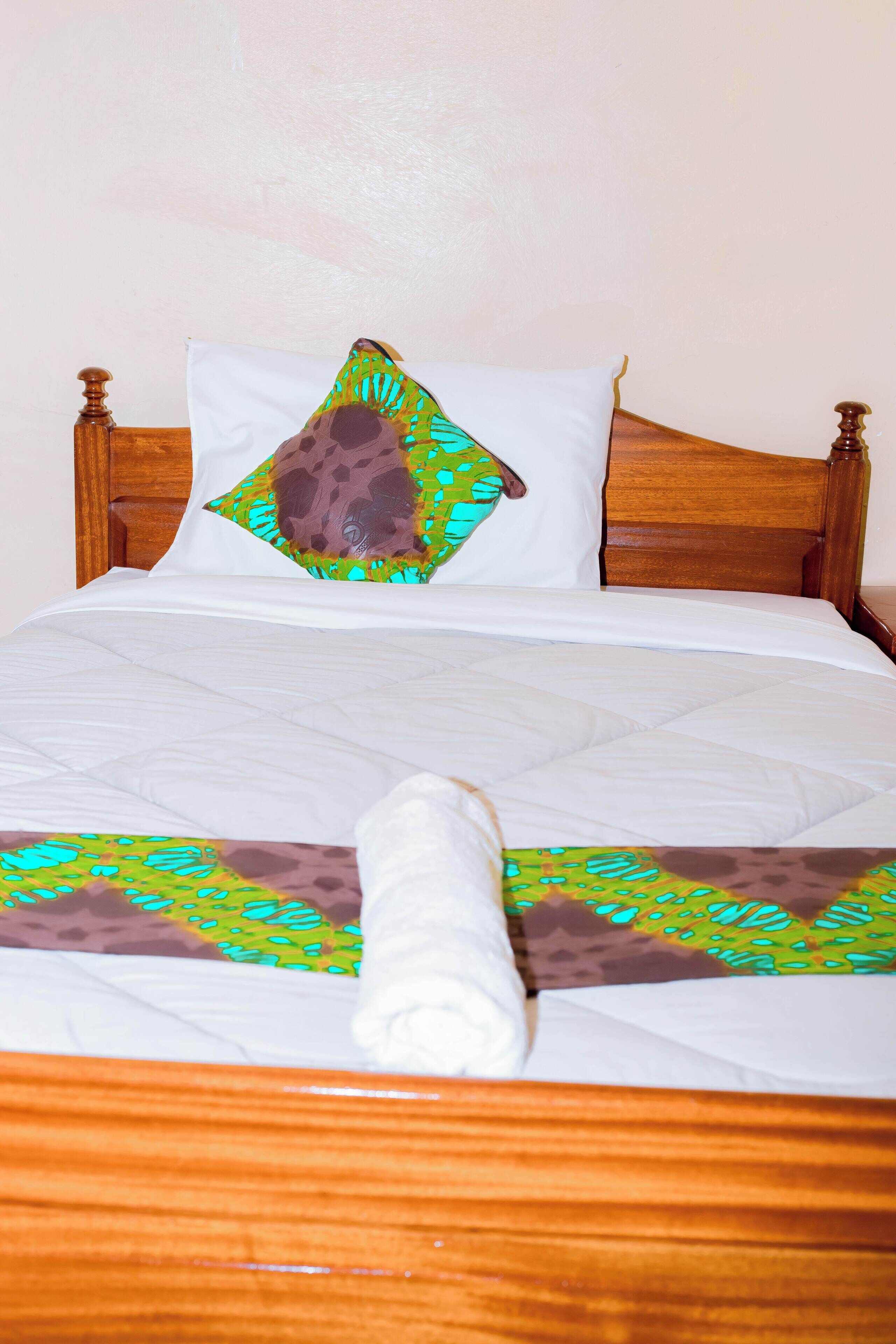 Una cama con cabecera de madera, almohada blanca y cojín decorativo con estampado verde y marrón.