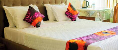 A neatly made bed with white linens and colorful pillows, a wooden headboard, and a table with a kettle and water bottle in the background.