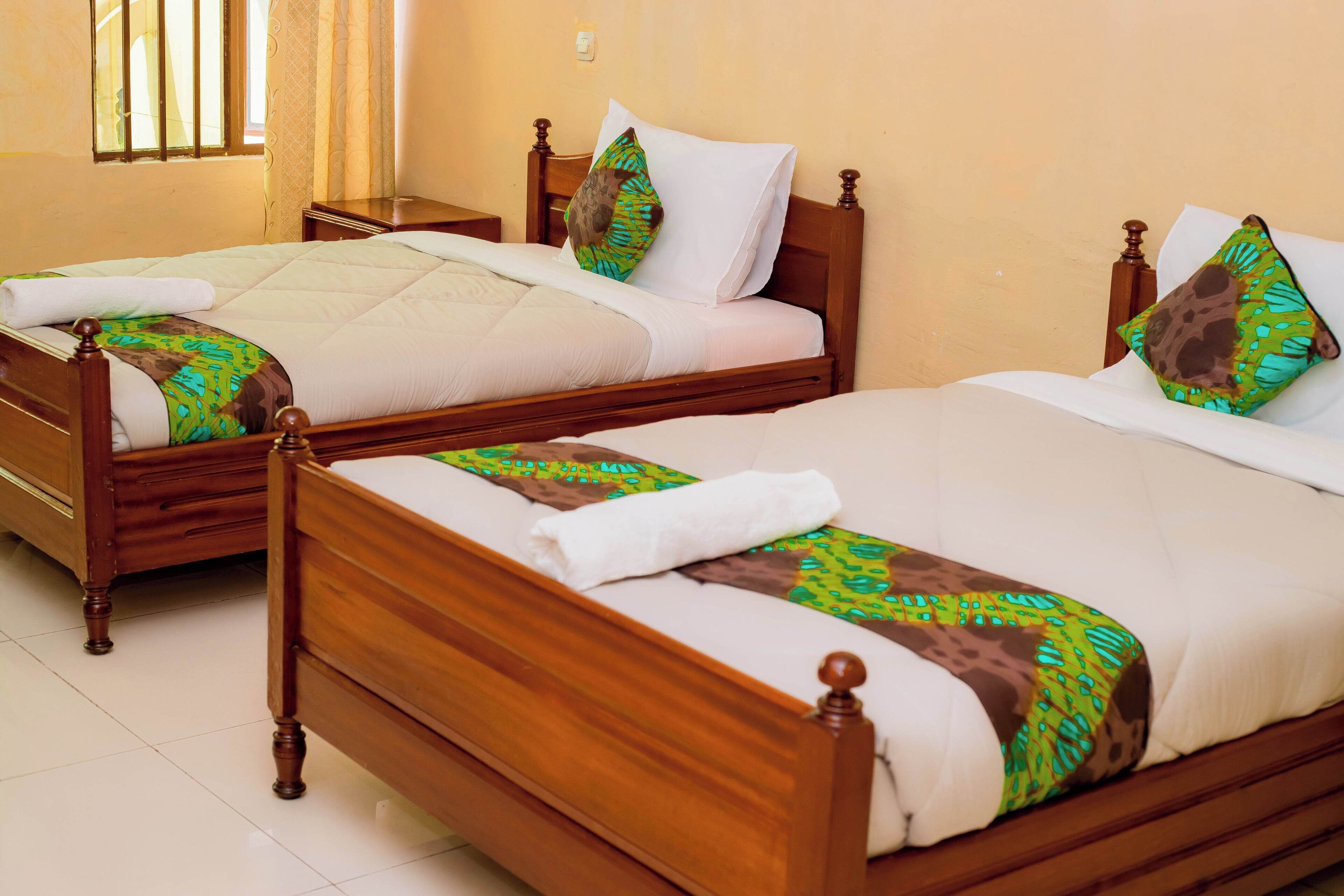 Two wooden beds with white linens and patterned pillows, in a room with a tiled floor and a window with bars.