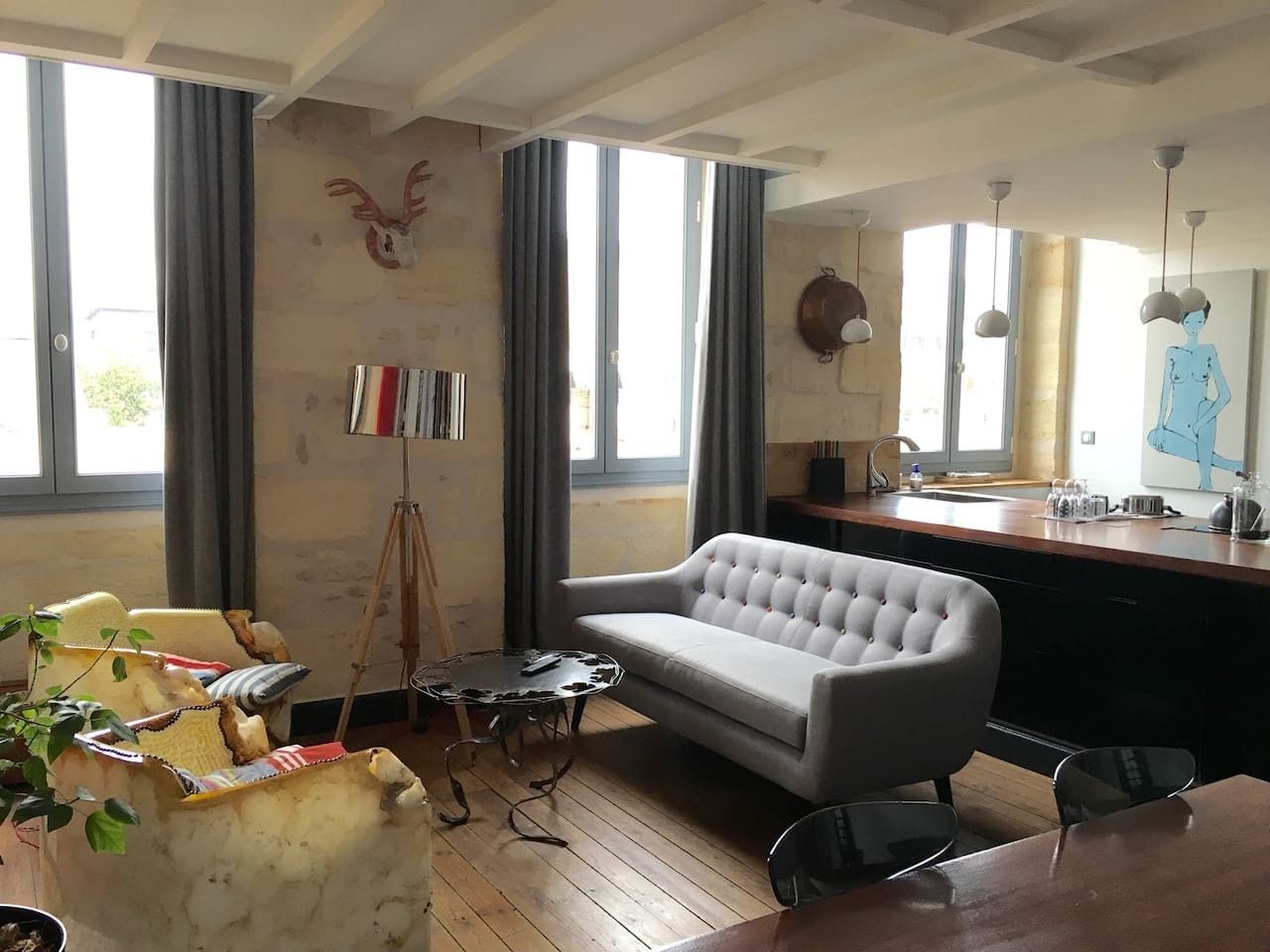 A living room with a sofa, a coffee table, and a dining table. There is a window with curtains, a wall-mounted sculpture, and a painting.