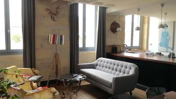 A living room with a sofa, a coffee table, and a dining table. There is a window with curtains, a wall-mounted sculpture, and a painting.