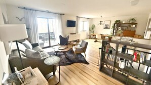 Ein geräumiger Wohnbereich mit einer Couch, Sesseln, einem Couchtisch und einem Esstisch. Ein Bücherregal mit Dekorationselementen und ein Flachbildfernseher an der Wand sind ebenfalls vorhanden.