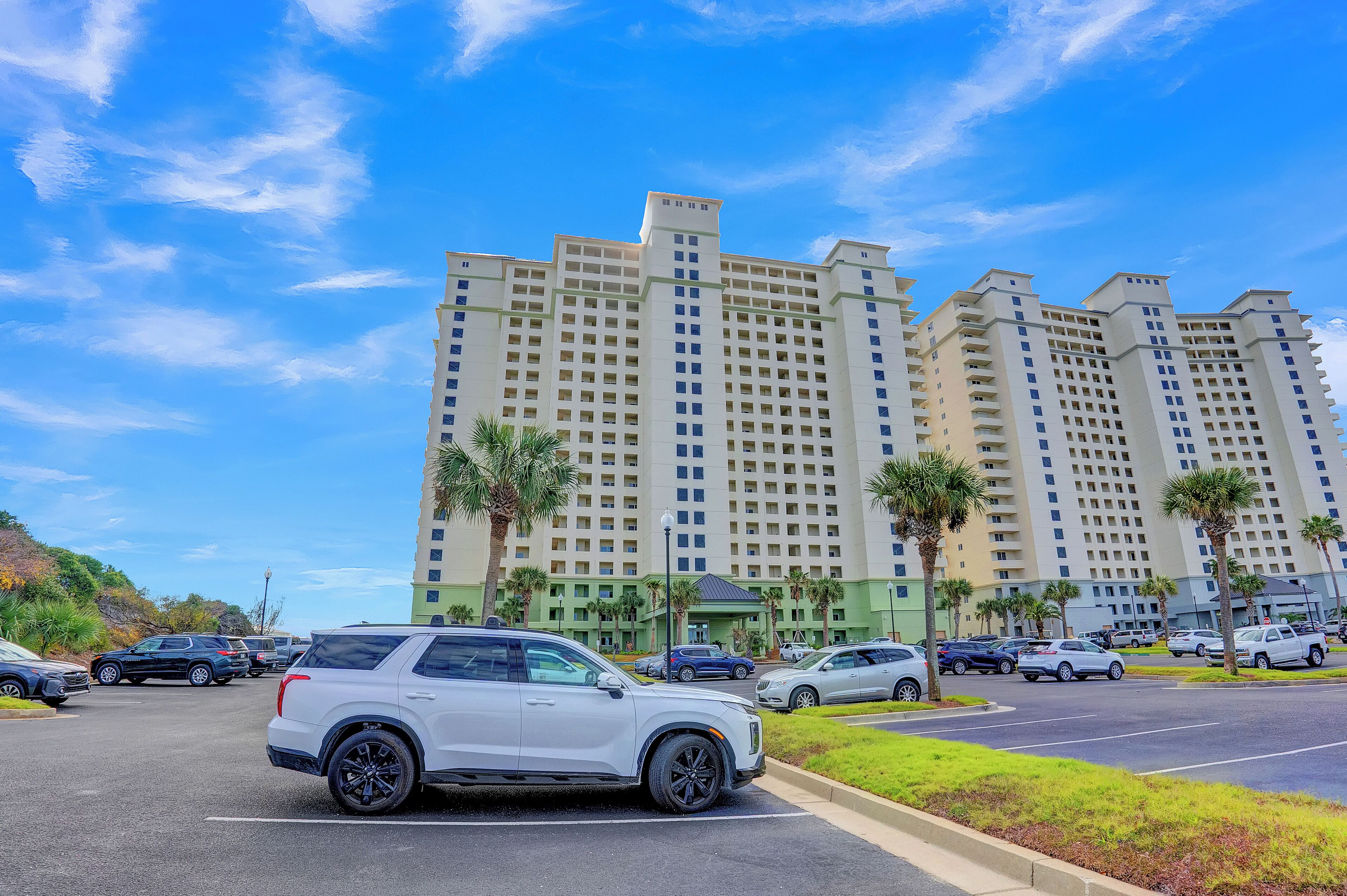 A multi-story hotel with a parking lot in front, featuring several cars and palm trees.