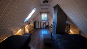 A cozy attic bedroom with wooden slats, two beds, a small table, and a window providing natural light.
